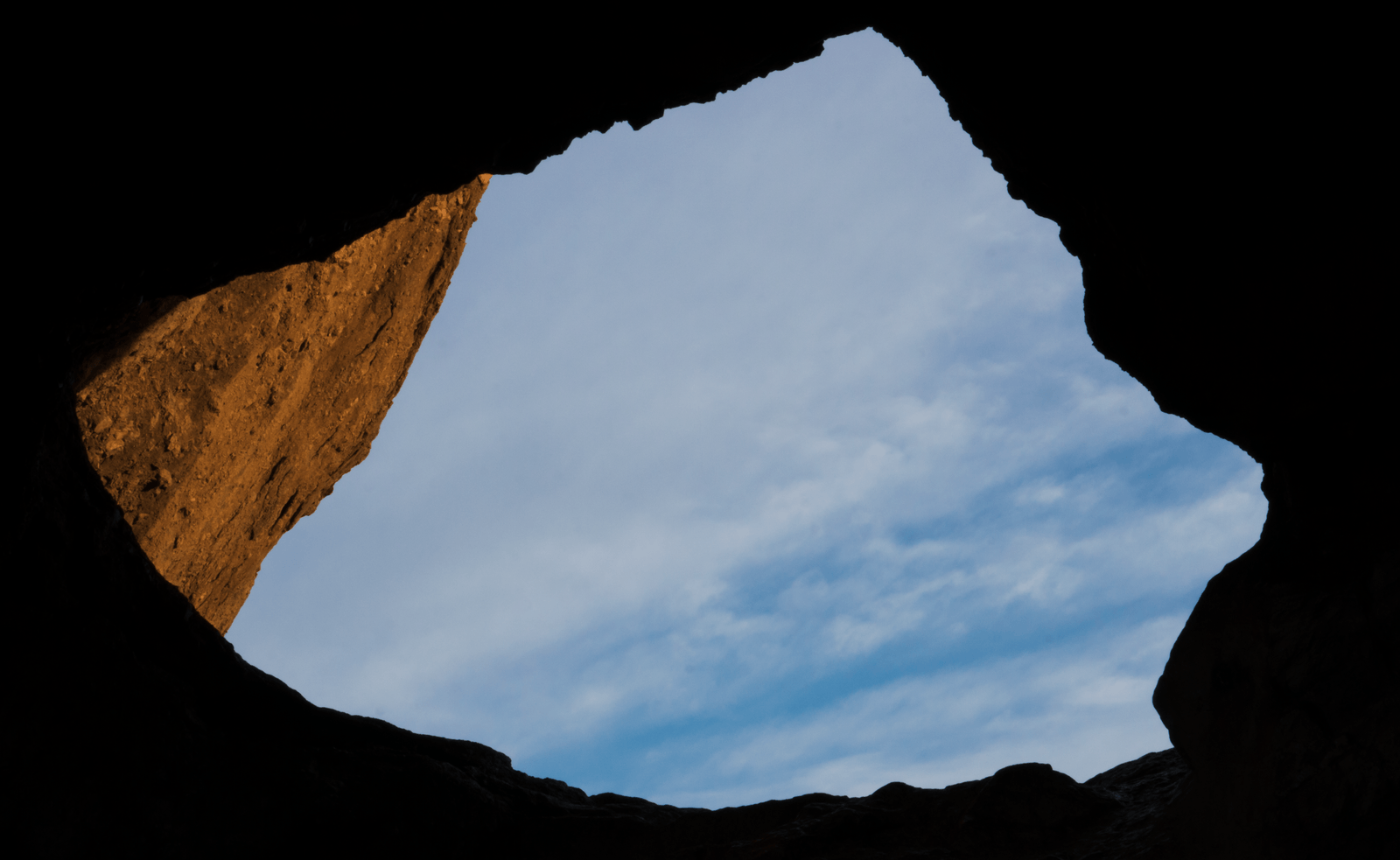 Looking up out of a cave mouth at a pale blue sky, with low sun on one rock wall.